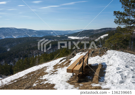 Gremyachaya Griva Park in Krasnoyarsk. Wooden bench on a snowy hill overlooking a winter forest valley. Peaceful mountain landscape for travel blog, nature magazine, outdoor tourism. Gremyachaya Griva Park in Krasnoyarsk. Wooden bench on a snowy hill overlooking a winter forest valley. Peaceful mountain landscape for travel blog, nature magazine, outdoor tourism. 136239665