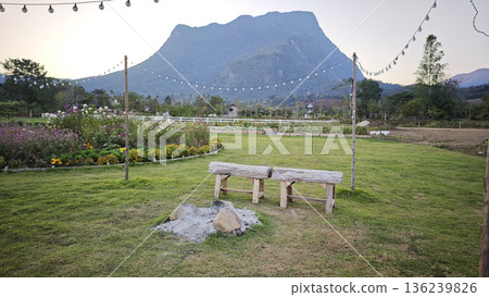 A beautifully landscaped flower garden with wooden tables for relaxing, offering a backdrop of mountains. A beautifully landscaped flower garden with wooden tables for relaxing, offering a backdrop of mountains. 136239826