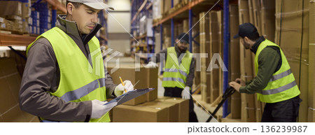 Warehouse workers doing inventory with clipboard while loader moves boxes on racks 136239987