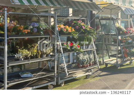 Vibrant outdoor flower shop display featuring colorful blooms, potted plants, and festive wreaths neatly arranged on tiered trolleys under striped awnings 136240117