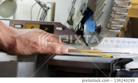 A close-up of a miter saw in action, a carpenter's hand holding a small round wooden dowel and sawing off a section on a miter saw with a sharp rotating blade A close-up of a miter saw in action, a carpenter's hand holding a small round wooden dowel and sawing off a section on a miter saw with a sharp rotating blade 136240299