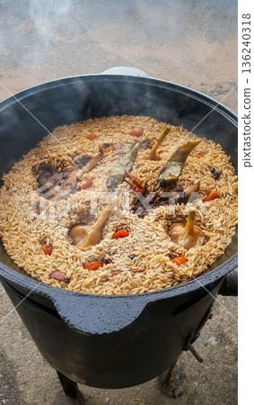 The photograph showcases a steaming pot of plov filled with tender meat and fresh vegetables, cooking over a flame. 136240318