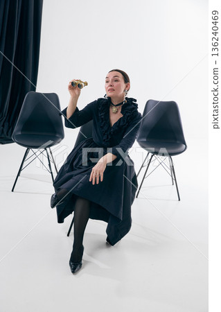 Elegant woman in black dress sitting with opera glasses in studio with curious expression 136240469