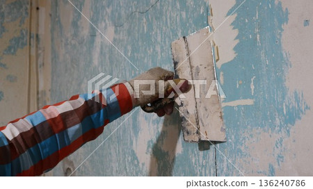scraping off the remains of wallpaper from peeling walls with a wide trowel, a close-up of a hand in a plaid shirt and a dirty glove with a spatula near the wall of a room, picking off scraps scraping off the remains of wallpaper from peeling walls with a wide trowel, a close-up of a hand in a plaid shirt and a dirty glove with a spatula near the wall of a room, picking off scraps 136240786