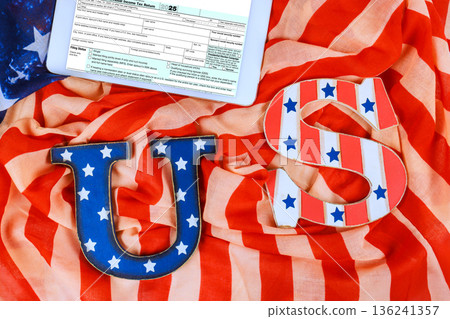 Letters US are placed on American flag with blue stars 1040 tax form digital tablet sit nearby. Letters US are placed on American flag with blue stars 1040 tax form digital tablet sit nearby. 136241357
