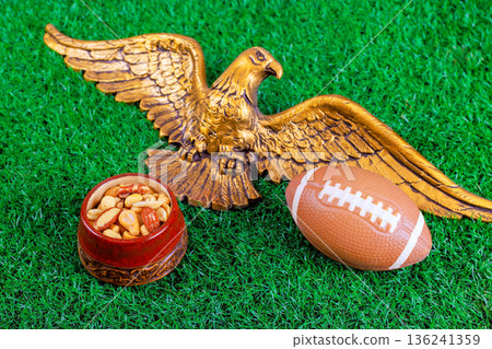 An eagle statue is placed bowl of mixed nuts American football on green grass for super bowl sports gathering. An eagle statue is placed bowl of mixed nuts American football on green grass for super bowl sports gathering. 136241359