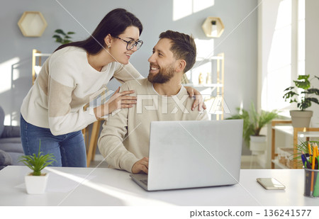 Happy loving woman looking at her husband while he is sitting at his working desk with his laptop Happy loving woman looking at her husband while he is sitting at his working desk with his laptop 136241577