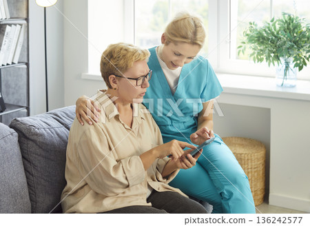 Kind caregiver nurse and senior woman sitting on sofa in home, older patient pointing at smartphone Kind caregiver nurse and senior woman sitting on sofa in home, older patient pointing at smartphone 136242577