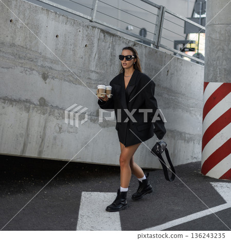 Woman in oversized black blazer and sunglasses holding takeaway coffee cups, walking in city street with confidence and style. Beautiful urban lady with coffee near concrete wall Woman in oversized black blazer and sunglasses holding takeaway coffee cups, walking in city street with confidence and style. Beautiful urban lady with coffee near concrete wall 136243235