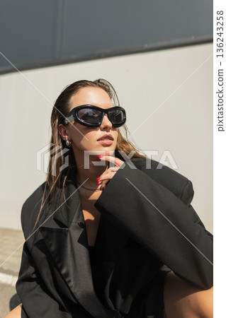 Beautiful cool urban young woman model with glamorous sunglasses in fashion black oversized blazer posing outdoors in the sun rays. Stylish trendy lady Beautiful cool urban young woman model with glamorous sunglasses in fashion black oversized blazer posing outdoors in the sun rays. Stylish trendy lady 136243258
