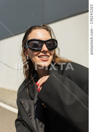 Stylish model in oversized blazer and shades enjoying sunny day, posing with a playful gesture, perfect for lifestyle, streetwear, and fashion concepts. 136243263