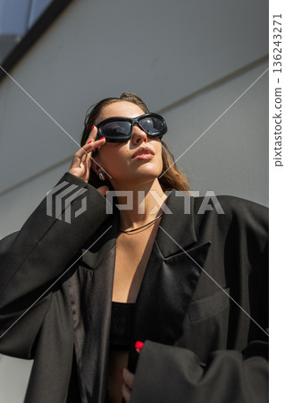 Young female model in trendy outfit with oversized jacket and accessories, posing outdoors in sunlight. Cool beautiful girl in black clothes puts on sunglasses in the city 136243271