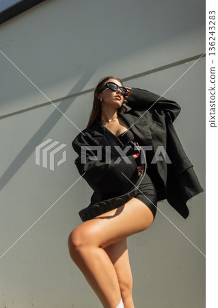 Stylish woman in oversized black blazer and sunglasses posing outdoors against modern architecture. Trendy street fashion and urban lifestyle concept. 136243283