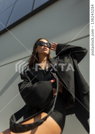 Fashionable young woman in oversized black blazer and sunglasses posing outdoors under sunlight. Modern street style, elegance, confidence, and trendy urban lifestyle concept. 136243284