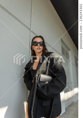 Stylish model in oversized jacket, boots, and sunglasses walking outdoors, perfect example of modern streetwear look. Fashionable black handbag 136243302