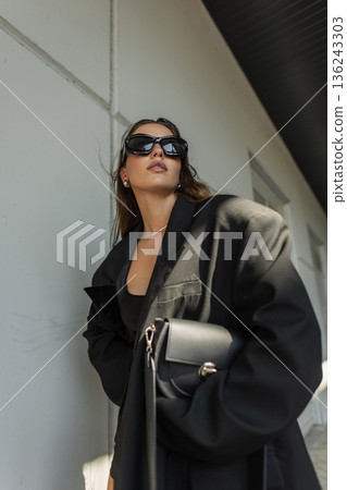 Confident young woman in oversized blazer with wet hair look, stylish sunglasses and natural makeup, modern street fashion portrait. Fashionable black handbag 136243303