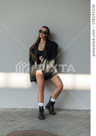 Stylish young woman in oversized black blazer, crop top, and boots posing against a gray wall, showcasing modern street fashion and confident attitude in natural light. 136243326