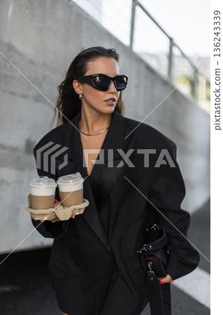 Stylish girl with bold sunglasses and black blazer holding two takeaway coffees, walking confidently in urban environment. Beautiful urban lady with coffee near concrete wall Stylish girl with bold sunglasses and black blazer holding two takeaway coffees, walking confidently in urban environment. Beautiful urban lady with coffee near concrete wall 136243339