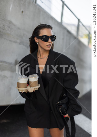 Trendy female in street style outfit, oversized blazer and accessories, holding coffee cups, urban lifestyle photography. Beautiful urban lady with coffee near concrete wall Trendy female in street style outfit, oversized blazer and accessories, holding coffee cups, urban lifestyle photography. Beautiful urban lady with coffee near concrete wall 136243341