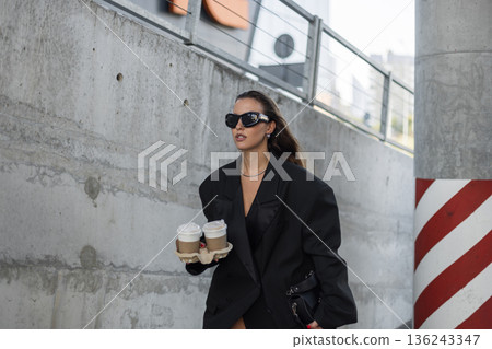 Fashionable woman in oversized black blazer and sunglasses carrying takeaway coffee cups, walking confidently in urban street setting. Fashionable woman in oversized black blazer and sunglasses carrying takeaway coffee cups, walking confidently in urban street setting. 136243347