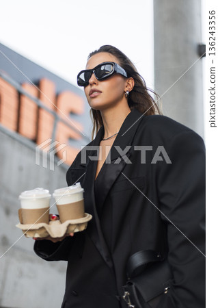 Elegant woman with wet hair look, chic black blazer and bold sunglasses, carrying two takeaway coffees in the city. Beautiful urban lady with coffee near concrete wall Elegant woman with wet hair look, chic black blazer and bold sunglasses, carrying two takeaway coffees in the city. Beautiful urban lady with coffee near concrete wall 136243356