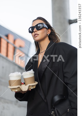 Stylish young female in trendy oversized blazer holding coffee to go, wearing black accessories and sunglasses, modern street lifestyle. Beautiful urban lady with coffee near concrete wall Stylish young female in trendy oversized blazer holding coffee to go, wearing black accessories and sunglasses, modern street lifestyle. Beautiful urban lady with coffee near concrete wall 136243357