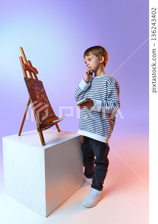 Child artist standing near easel and painting in studio. Child artist standing near easel and painting in studio. 136243402