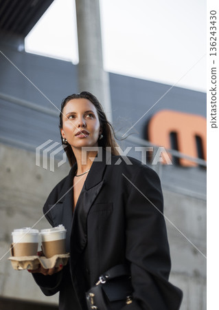 Trendy woman wearing oversized blazer carrying takeaway drinks, showcasing minimalist fashion and contemporary urban lifestyle. Trendy woman wearing oversized blazer carrying takeaway drinks, showcasing minimalist fashion and contemporary urban lifestyle. 136243430
