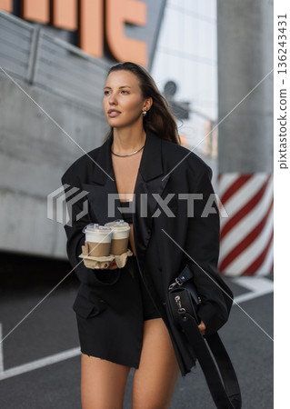 Fashionable woman in black outfit with coffee to go, walking confidently in the urban environment, symbolizing street style and city elegance. Fashionable woman in black outfit with coffee to go, walking confidently in the urban environment, symbolizing street style and city elegance. 136243431