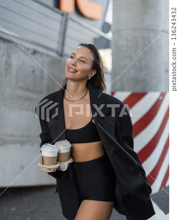 Smiling young woman in black blazer and crop top holding takeaway coffee cups outdoors, showing modern street fashion, confidence, and active urban lifestyle. Smiling young woman in black blazer and crop top holding takeaway coffee cups outdoors, showing modern street fashion, confidence, and active urban lifestyle. 136243432