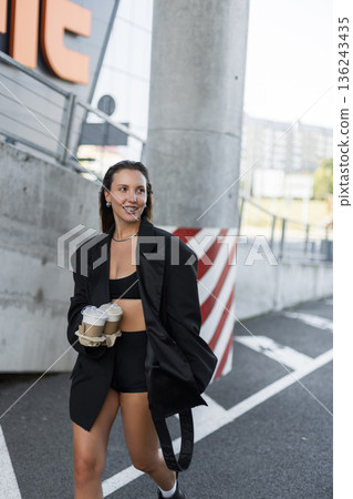 Cheerful young woman in black stylish outfit carrying takeaway coffee cups outdoors, enjoying a casual walk in the city with a bright smile and confident street style. Cheerful young woman in black stylish outfit carrying takeaway coffee cups outdoors, enjoying a casual walk in the city with a bright smile and confident street style. 136243435