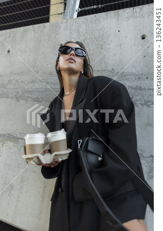 Stylish woman in oversized black blazer and sunglasses holding takeaway coffee cups, posing against a concrete wall in modern urban street style. Stylish woman in oversized black blazer and sunglasses holding takeaway coffee cups, posing against a concrete wall in modern urban street style. 136243451