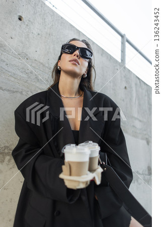 Stylish woman in oversized black blazer and sunglasses carrying coffee cups, stands near concrete wall in the city. Pretty business urban lady Stylish woman in oversized black blazer and sunglasses carrying coffee cups, stands near concrete wall in the city. Pretty business urban lady 136243452