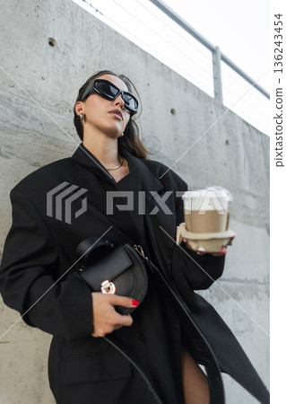 Confident woman in fashionable street style, carrying coffee and wearing oversized blazer with sunglasses stands near concrete wall. 136243454