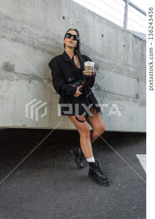 Fashionable woman in oversized black blazer and boots holding takeaway coffee, leaning against a concrete wall in urban city street style. Fashionable woman in oversized black blazer and boots holding takeaway coffee, leaning against a concrete wall in urban city street style. 136243456
