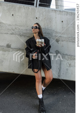 Stylish young woman in sunglasses and black outfit with coffee to go, standing outdoors by a concrete wall. Elegant young woman walking outside with takeaway coffee, dressed in stylish oversized black 136243457