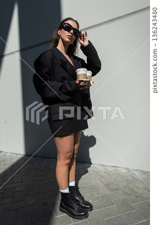 Stylish girl in black boots and sunglasses carrying coffee cups, posing against city wall in sunlight, trendy street fashion look. Stylish girl in black boots and sunglasses carrying coffee cups, posing against city wall in sunlight, trendy street fashion look. 136243480