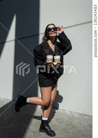 Fashionable young woman in oversized black jacket, sunglasses and boots holding takeaway coffee cups outdoors, urban street style, modern lifestyle and casual confidence. Fashionable young woman in oversized black jacket, sunglasses and boots holding takeaway coffee cups outdoors, urban street style, modern lifestyle and casual confidence. 136243483