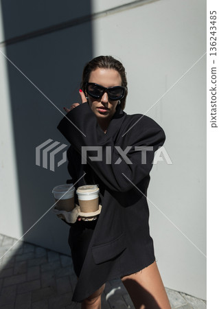 Fashionable woman in oversized black blazer and boots holding two takeaway coffee cups, posing against a sunlit wall. Trendy street style and modern urban lifestyle concept. Fashionable woman in oversized black blazer and boots holding two takeaway coffee cups, posing against a sunlit wall. Trendy street style and modern urban lifestyle concept. 136243485
