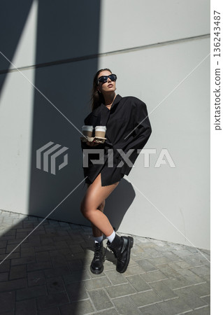 Stylish woman in black oversized blazer, sunglasses and boots holding takeaway coffee cups, walking outdoors on sunny day, modern street fashion and urban lifestyle concept. Stylish woman in black oversized blazer, sunglasses and boots holding takeaway coffee cups, walking outdoors on sunny day, modern street fashion and urban lifestyle concept. 136243487