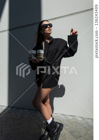 Stylish woman with long hair wearing black blazer and sunglasses, carrying coffee cups, modern city lifestyle and streetwear look. Stylish woman with long hair wearing black blazer and sunglasses, carrying coffee cups, modern city lifestyle and streetwear look. 136243489