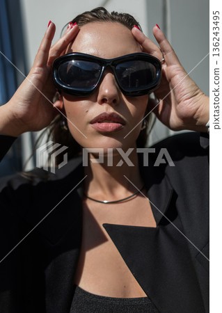 Close-up portrait of a stylish woman in oversized black sunglasses and blazer, posing confidently outdoors in natural sunlight, showcasing modern fashion and urban elegance. Close-up portrait of a stylish woman in oversized black sunglasses and blazer, posing confidently outdoors in natural sunlight, showcasing modern fashion and urban elegance. 136243495