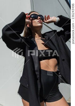 Cool urban beautiful woman model in fashionable black clothes with blazer wears glasses on sunny day in city. Sexy pretty lady outdoors 136243496
