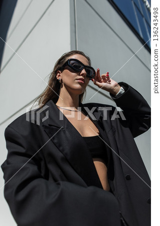 urban beautiful fashion woman in stylish black oversized blazer putting on sunglasses and walking in city on sunny day. Pretty glam girl urban beautiful fashion woman in stylish black oversized blazer putting on sunglasses and walking in city on sunny day. Pretty glam girl 136243498