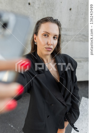 Confident woman in oversized black blazer taking a selfie with smartphone outdoors, representing modern fashion, technology, and urban lifestyle. Confident woman in oversized black blazer taking a selfie with smartphone outdoors, representing modern fashion, technology, and urban lifestyle. 136243499