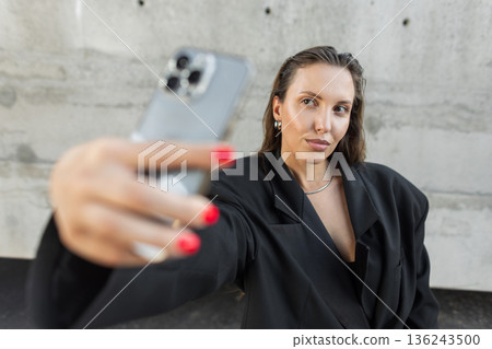 Stylish young woman in oversized black blazer taking a selfie with smartphone outdoors, showcasing modern street fashion and confident urban lifestyle. Stylish young woman in oversized black blazer taking a selfie with smartphone outdoors, showcasing modern street fashion and confident urban lifestyle. 136243500