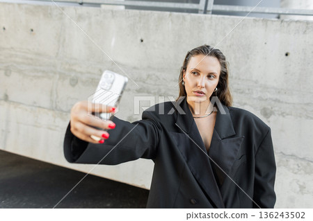 Stylish young woman in black oversized blazer posing for a selfie with smartphone against concrete wall, showcasing modern street style and confidence. Stylish young woman in black oversized blazer posing for a selfie with smartphone against concrete wall, showcasing modern street style and confidence. 136243502