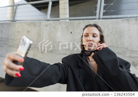 Confident fashion model in oversized blazer and dark sunglasses holding smartphone for a selfie, representing elegance, bold style, and city vibes. Confident fashion model in oversized blazer and dark sunglasses holding smartphone for a selfie, representing elegance, bold style, and city vibes. 136243504