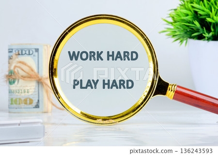 A magnifying glass focuses on the motivational phrase Work Hard Play Hard. Stacked US Dollar bills and a green plant are visible on a bright white desk A magnifying glass focuses on the motivational phrase Work Hard Play Hard. Stacked US Dollar bills and a green plant are visible on a bright white desk 136243593