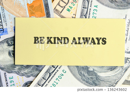A yellow note card with the inspiring words BE KIND ALWAYS rests on a bed of United States dollar bills, clearly showing Benjamin Franklin portraits 136243602